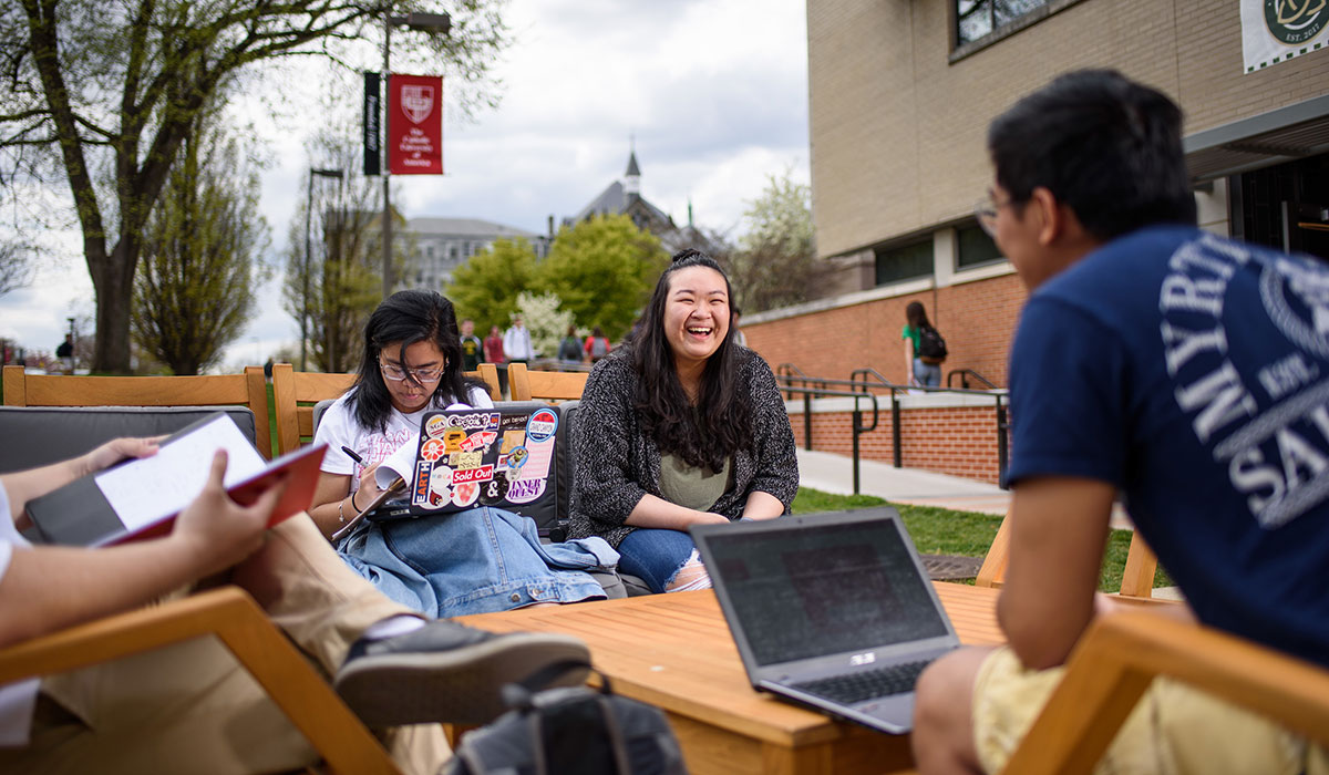 students laugh and talk at a table and chairs outside