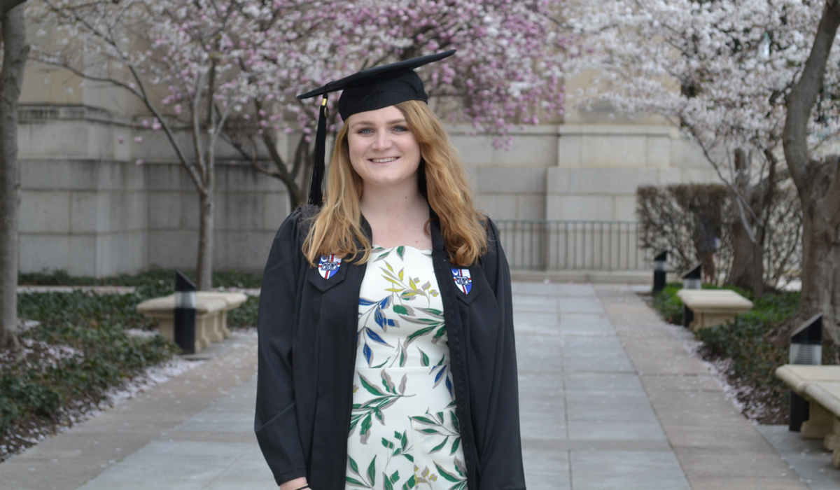 Colleen Yeckley standing in graduation gown under blossoming trees