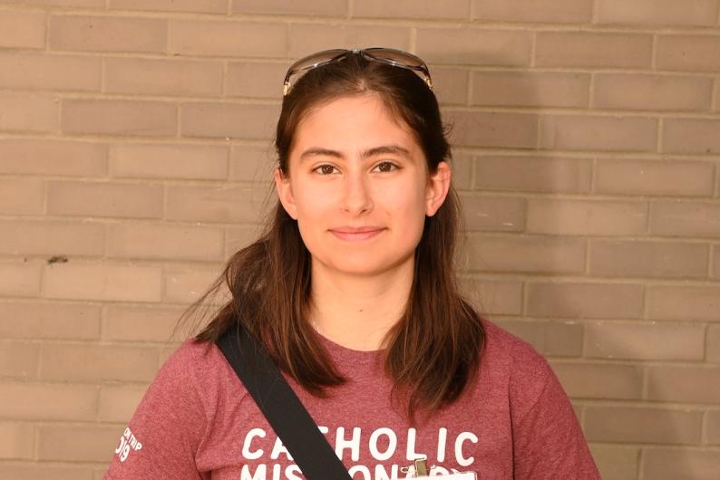 A woman with shoulder-length brown hair, wearing a Catholic University sweatshirt, stands in front of a brick wall.