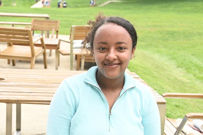 A woman is standing in front of a sprawling lawn darted with wooden chairs. Her curly hair is up in a bun and she is wearing a mint 3/4 zip sweater.