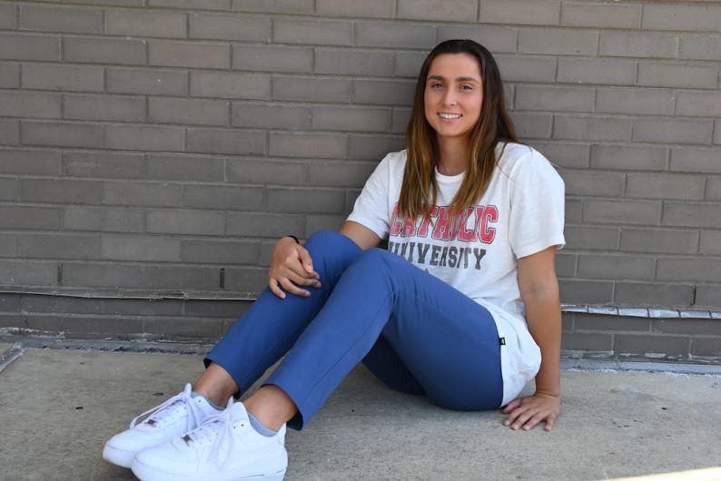 A woman sits on the ground and leans against a brick wall. She is wearing a Catholic University tee shirt, jeans, and white sneakers. She has long brown hair.