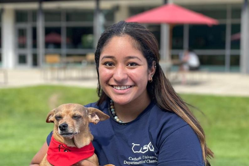 A woman with long brown hair and a blue shirt stands in front of the Pryz Center at Catholic University. She's holding a small dog wearing a red bandana.