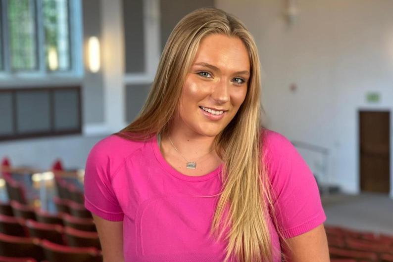A woman stands in the back of an auditorium. She is wearing a bright pink shirt and has long blonde hair. 