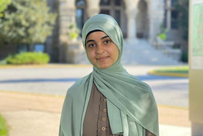 A woman stands in front of a court yard with a stone building and trees. She is wearing a grey dress and a mint green hijab