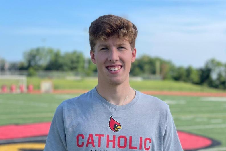 A man with curly blonde hair in a Catholic University shirt stands in front of a football field.
