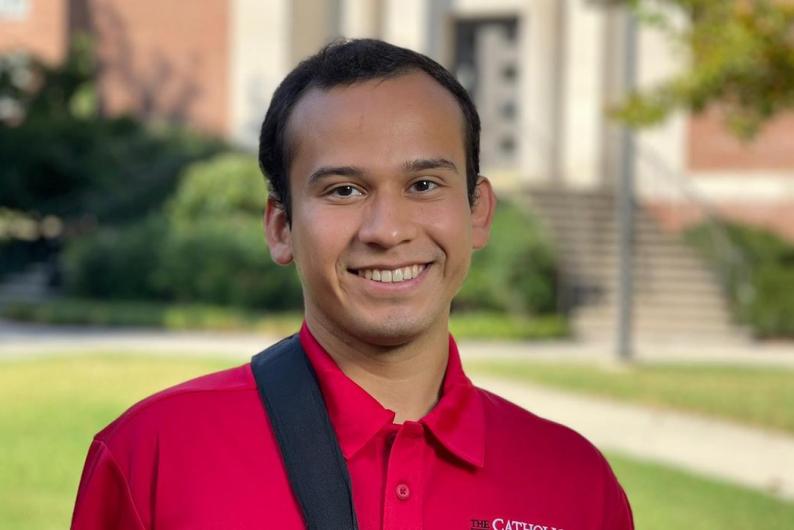A man is wearing a red polo in front of a red brick building, a bush, and a lush lawn. 