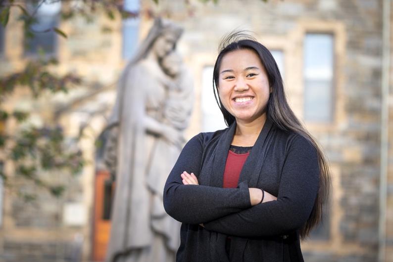 A woman with long black hair is standing with her arms crossed in front of a statue of Mary and Jesus and grey brick building. She is wearing a black cardigan and a red shirt.