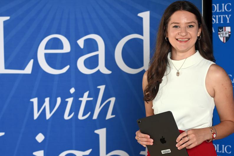 Julia Englert is standing in front of a Catholic University Lead With Light background. She has long brown hair and is wearing a white top and red pants. 