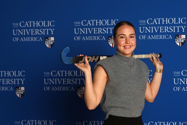 Mary Parker is standing in front of a blue Catholic University of America background. She has her field hockey stick draped over her shoulder.