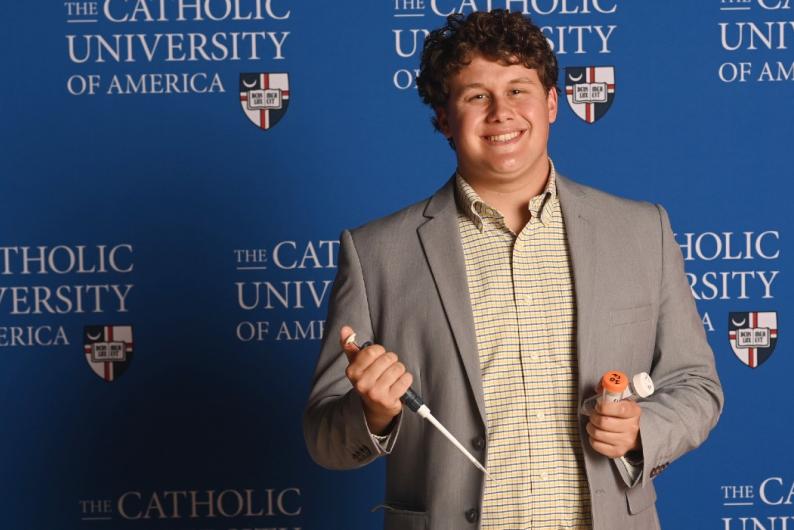 Elijah is standing in front of a Catholic University background. He has curly hair and is wearing a checkered shirt and a grey blazer.