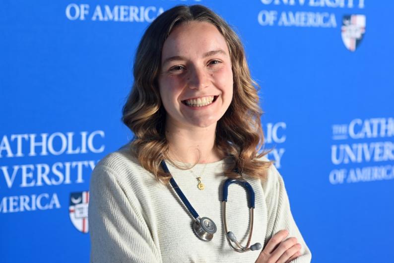 Elizabeth is standing in front of a blue Catholic University background. She is wearing white and has a stethoscope around her neck.