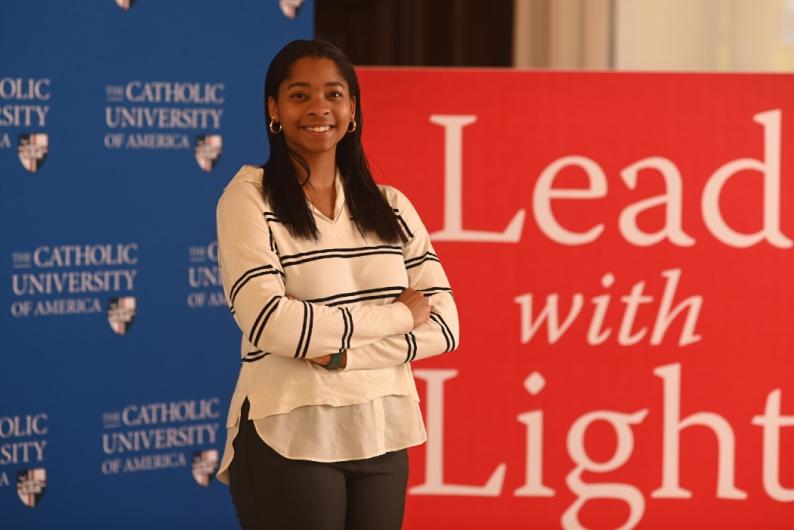 Lauren White is standing in front of blue and red Catholic University backdrops. Her arms are crossed and she's smiling. She is wearing a white and black sweater and dark pants.