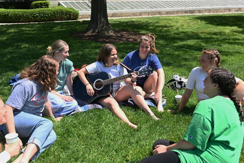 student plays the guitar on a lawn on campus with group of friends gathered around her