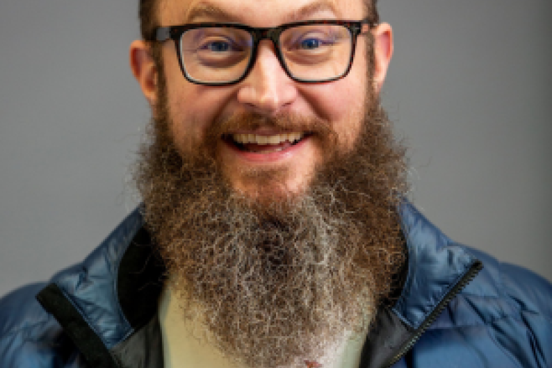 Taylor Black is a man with a long brown and grey beard. He has black, square glasses. He is wearing a grey shirt and a blue puffy vest.